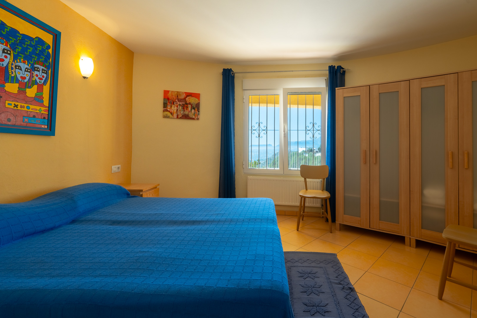 Bright bedroom with a double bed, ochre walls, a wooden wardrobe with translucent panels, and a window overlooking the coast.