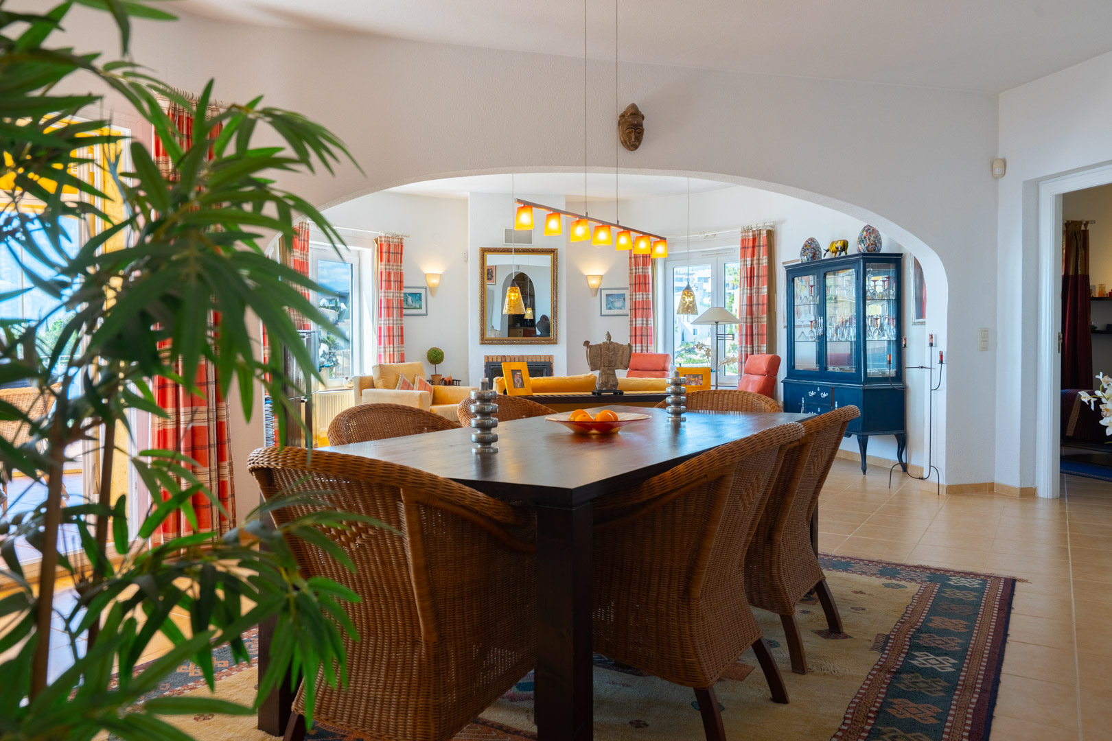 Bright dining area featuring a dark wood table and wicker chairs, connected to the living room via a wide archway. Tiled flooring and natural light.