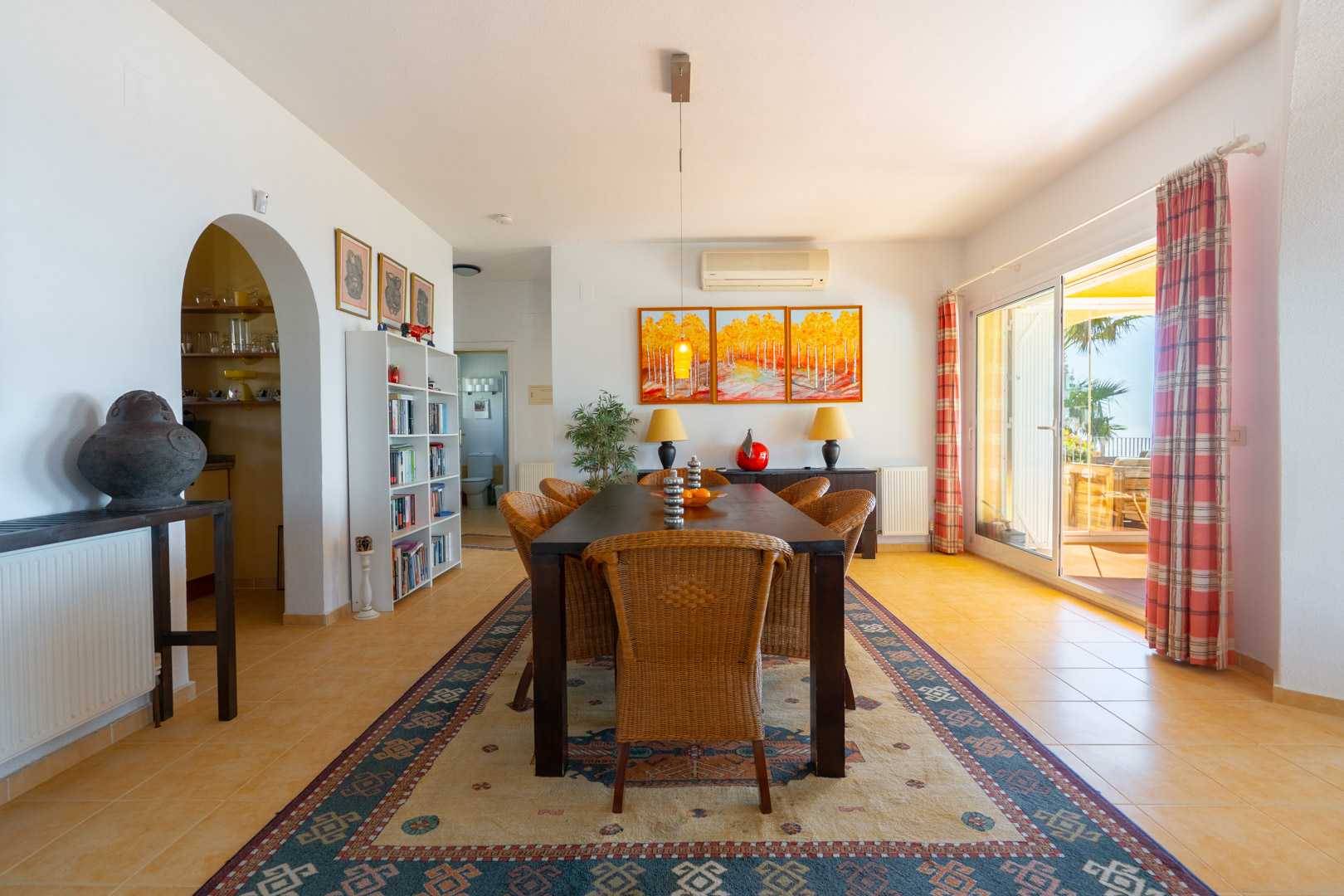 Bright dining area featuring a dark wood table with wicker chairs, traditional arched doorway, and large glass doors opening onto the sun terrace.