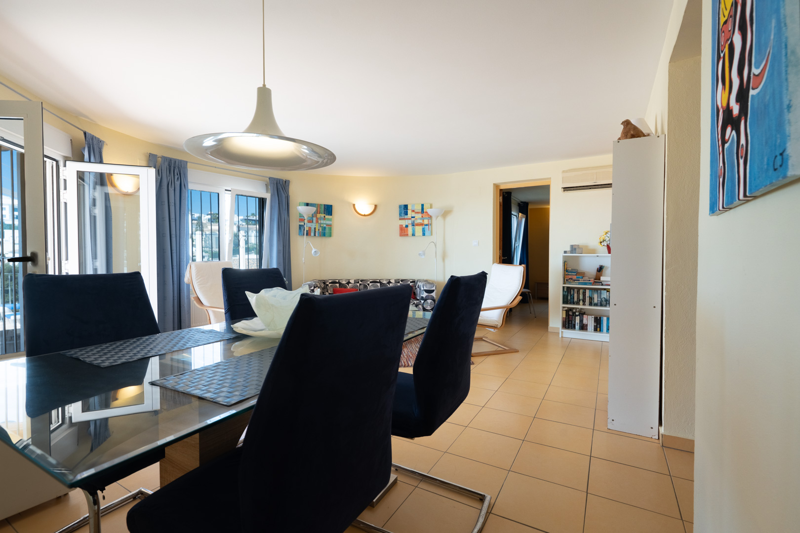 Bright dining area featuring a glass-top table and navy velvet chairs, set within an open-plan living space with ceramic tiling and natural light.