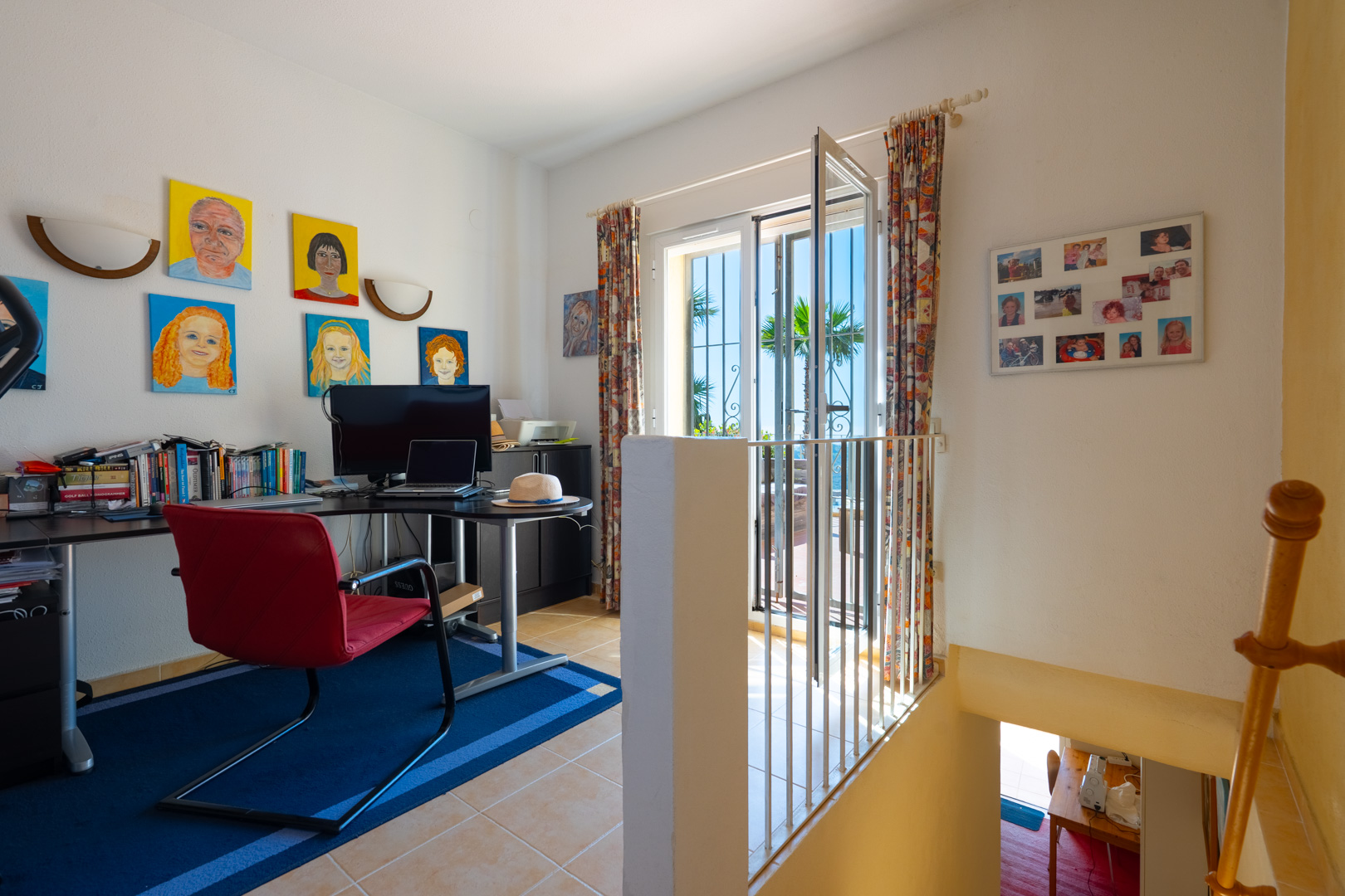 Bright home office featuring a corner desk, red chair, and terrace access. The space includes tiled flooring and the staircase leading to the lower level.