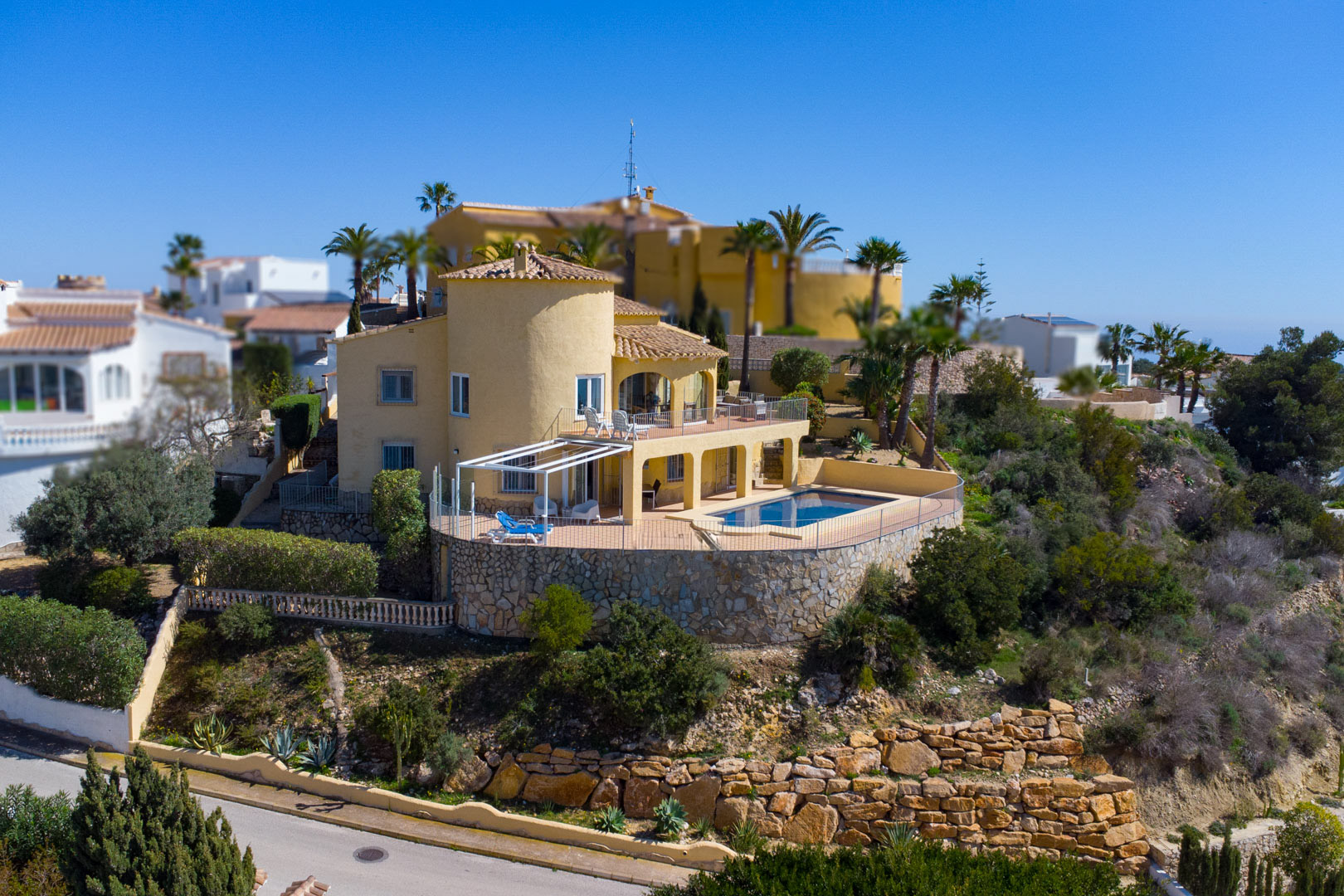 Exterior view of the villa featuring an ochre facade, a circular tower, and a rectangular swimming pool on a spacious terrace with stone retaining walls.