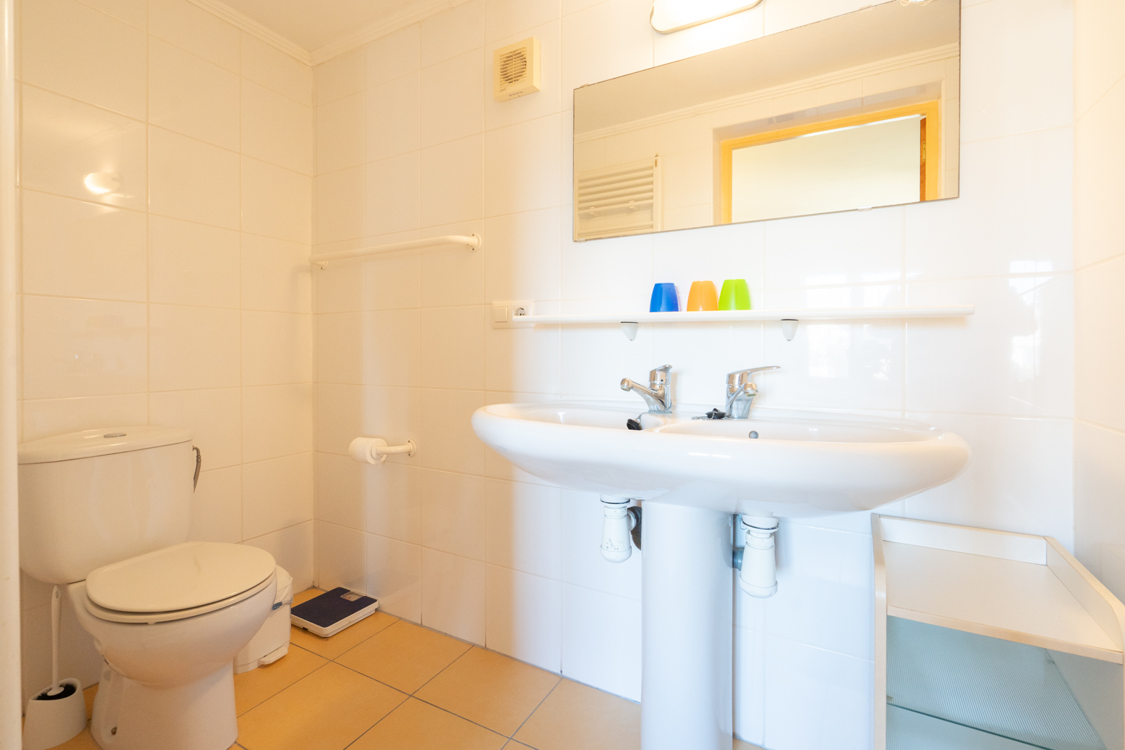 Functional bathroom featuring a double pedestal sink, white tiled walls, and warm-toned floor tiles. Equipped with a large mirror and integrated shelving.