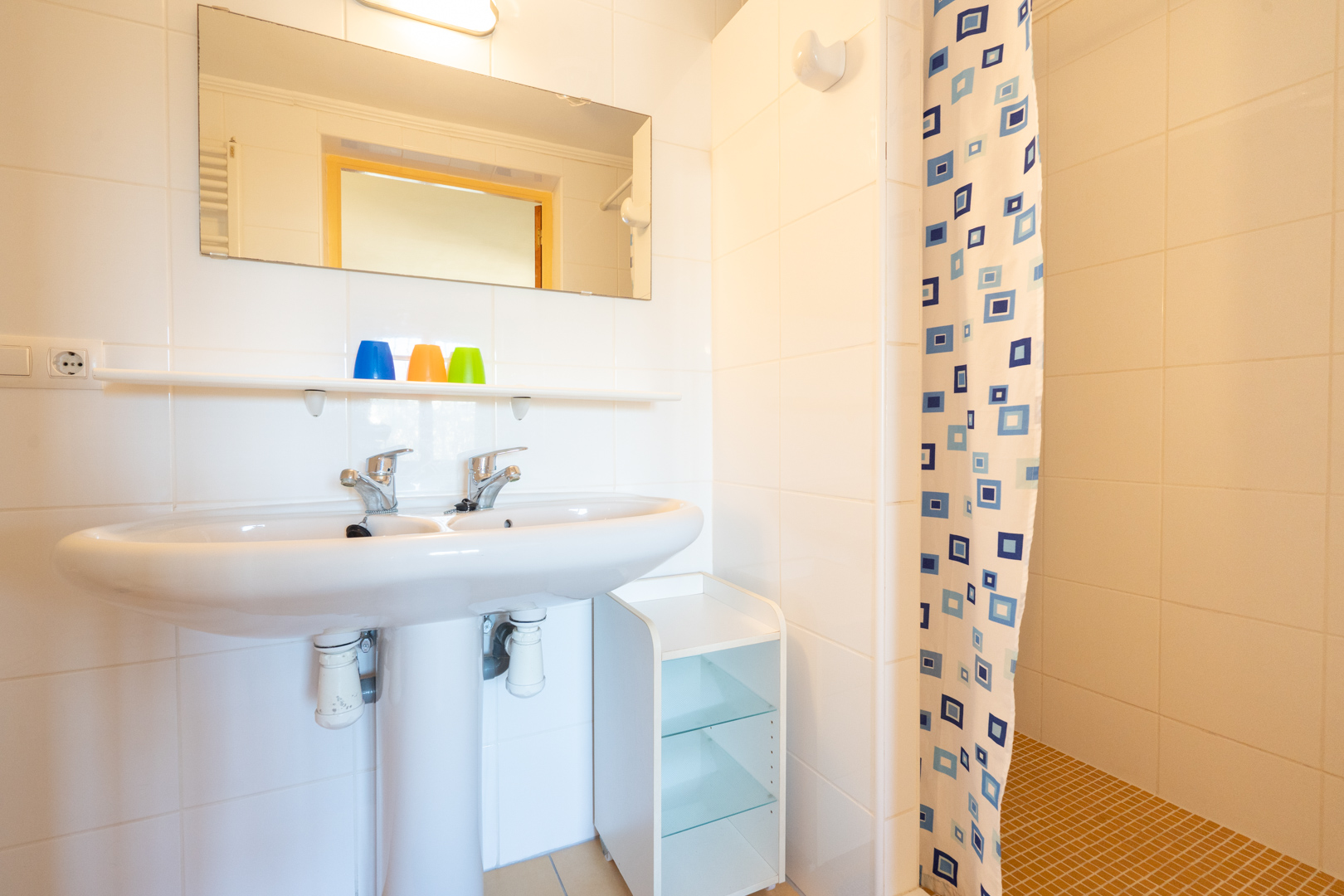 Functional bathroom featuring a double-tap pedestal sink, white tiled walls, and a shower area with a patterned curtain and ochre floor tiles.