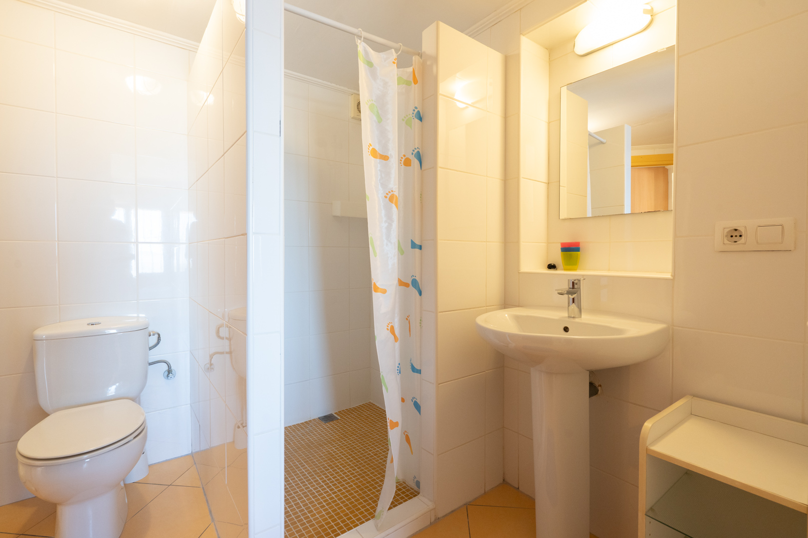 Functional bathroom featuring white wall tiles, a walk-in shower with a patterned curtain, a pedestal sink, and classic porcelain fixtures.