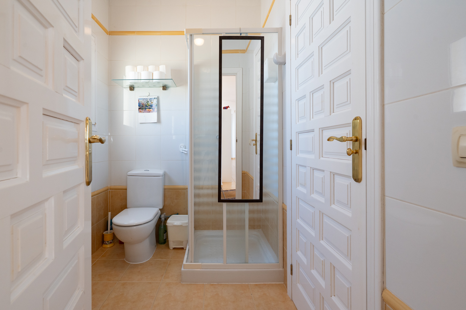 Functional bathroom with white and beige tiling, featuring a toilet and a shower cubicle with a mirror, framed by white paneled doors.