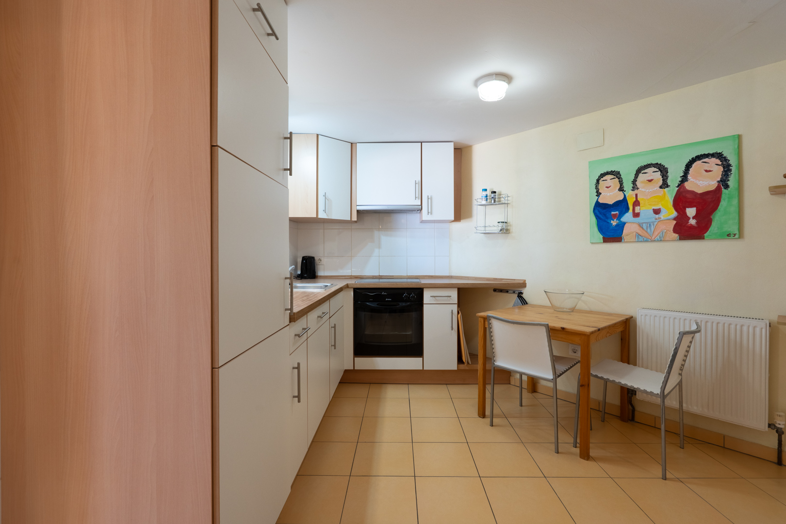 Functional guest apartment kitchen featuring white cabinetry, wood-effect countertops, a compact dining area, and warm-toned ceramic floor tiles.