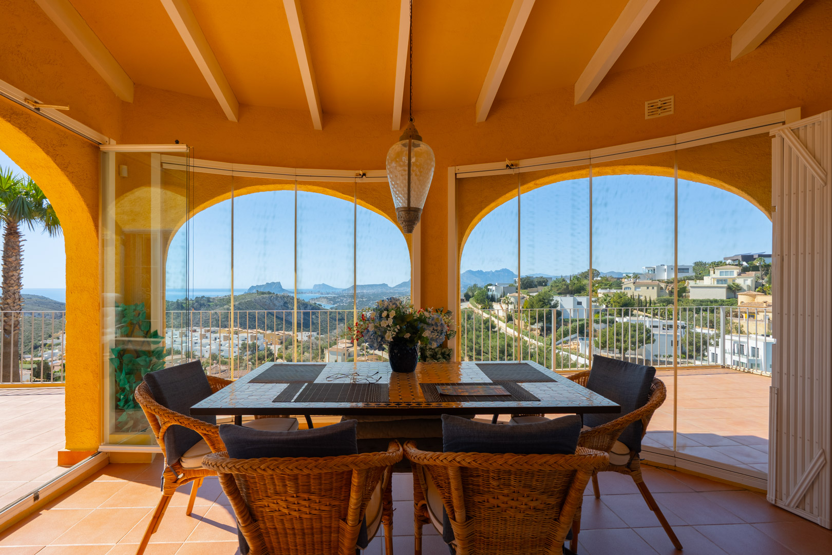 Glass-enclosed naya with traditional arches and wooden beams, offering panoramic sea and mountain views from the dining area with terracotta flooring.