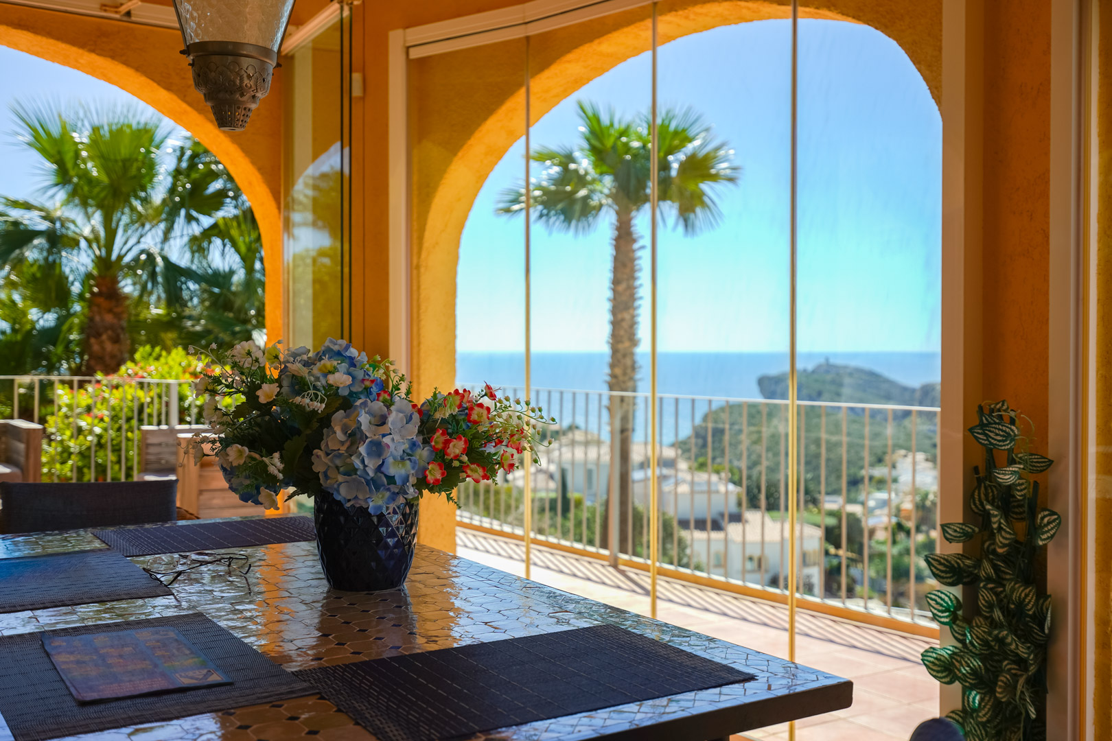 Glazed naya featuring a mosaic-tiled table and arched openings with glass curtains, showcasing panoramic views of the Mediterranean Sea and Cap d’Or.