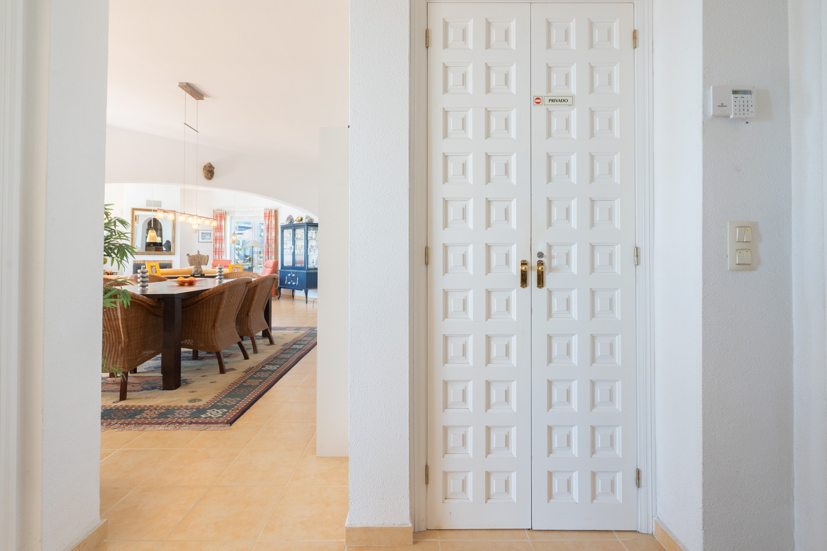 Interior hallway featuring white paneled double doors and a view into the dining area with wicker seating and traditional tiled flooring.