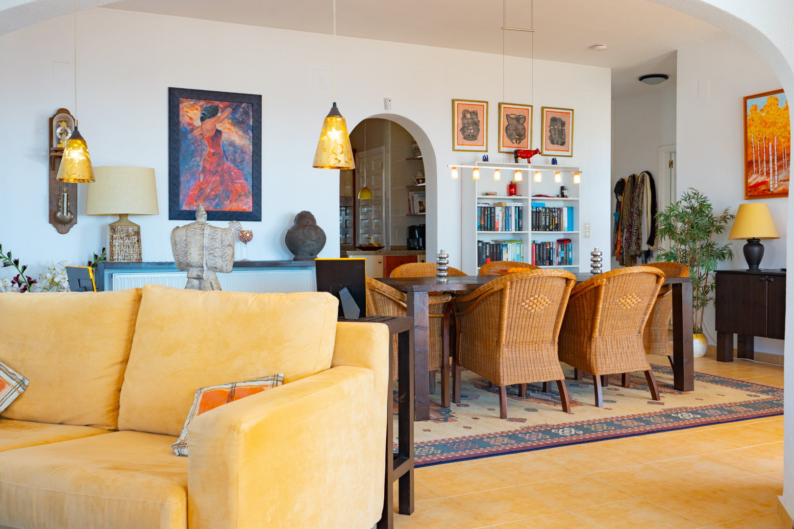 Open-plan living and dining space featuring wicker chairs, a dark wood table, and terracotta tiles. Warm pendant lighting and arched transitions define the interior layout.