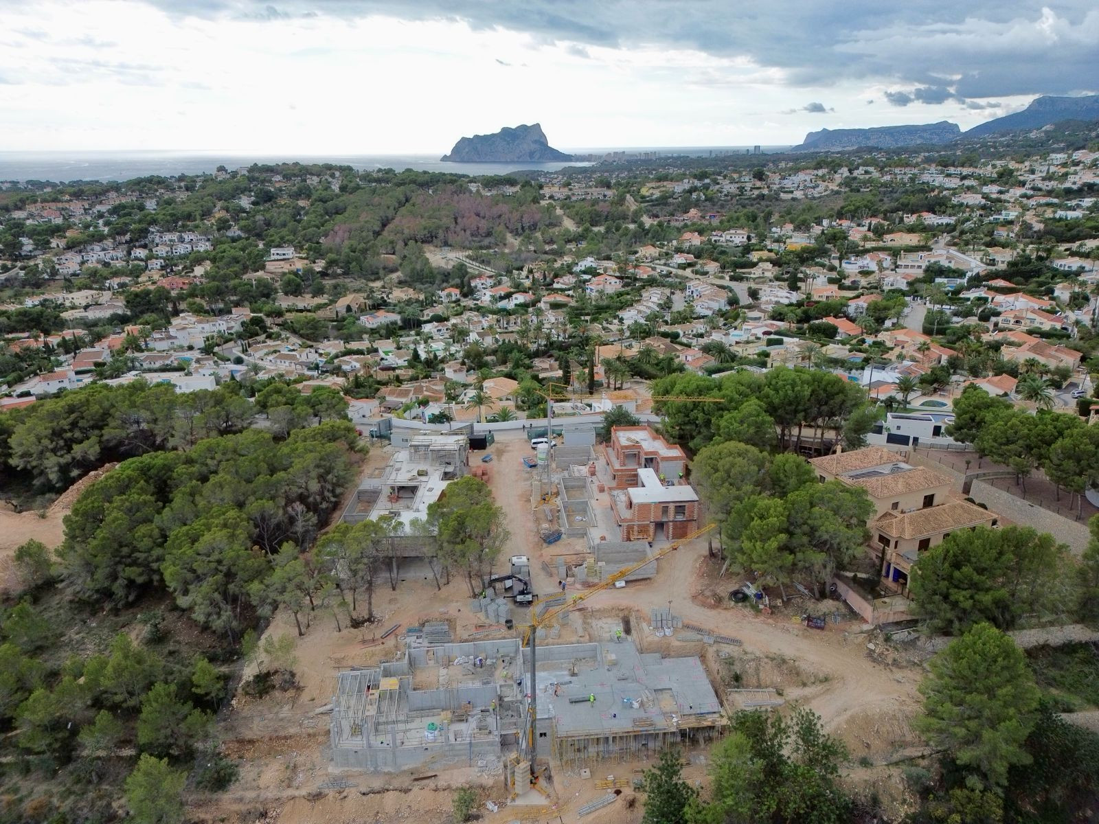 Aerial view of a villa construction site with concrete foundations and partially built walls, set within a residential area offering distant sea and mountain panoramas.