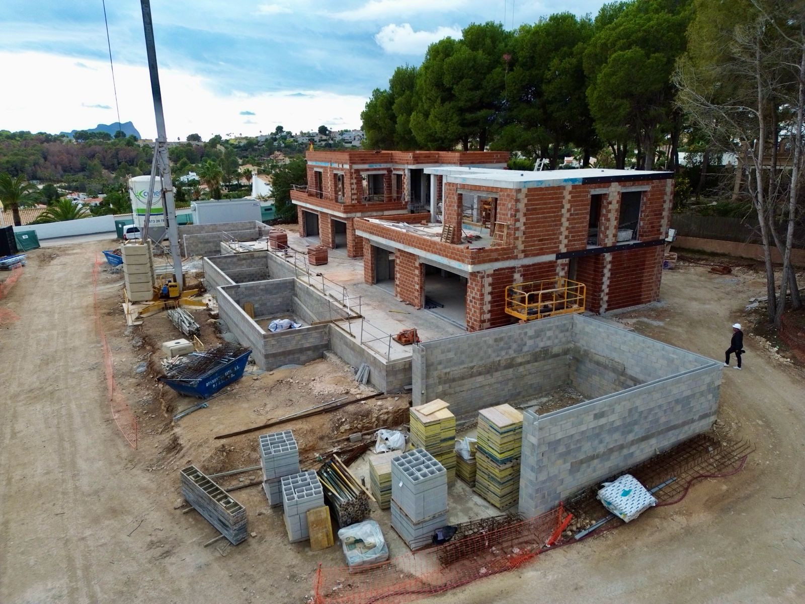 Aerial view of the villa construction site, featuring multiple brick structures in progress, excavated concrete foundations, and building materials. A crane is visible, with distant green hills.