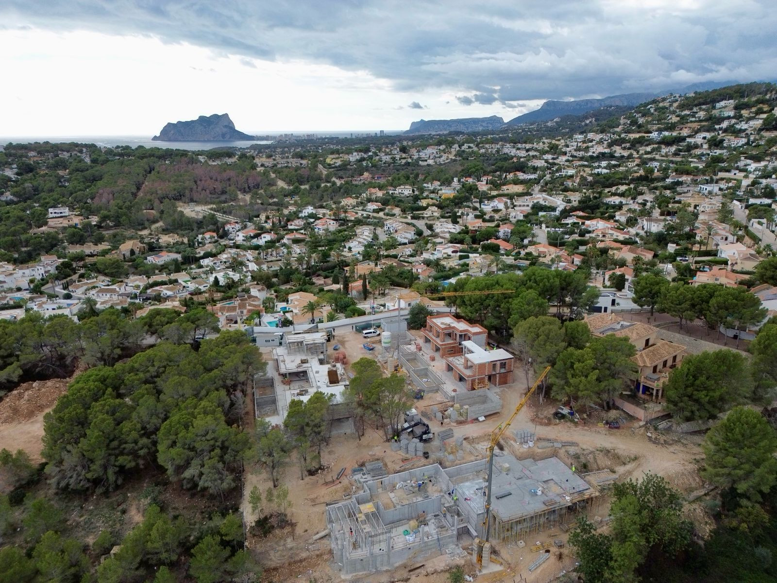 Aerial view of villas under construction within a residential area of Moraira, showing cranes and foundations, framed by the sea and Peñón de Ifach in the distance.