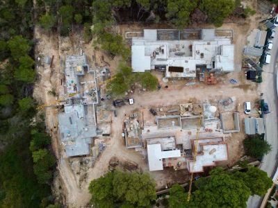 Aerial view of a villa complex under construction, featuring foundations, concrete slabs, and two cranes. Building materials and vehicles are visible on the site.