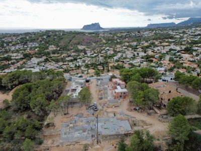 Aerial view of a villa construction site with concrete foundations and partially built walls, set within a residential area offering distant sea and mountain panoramas.