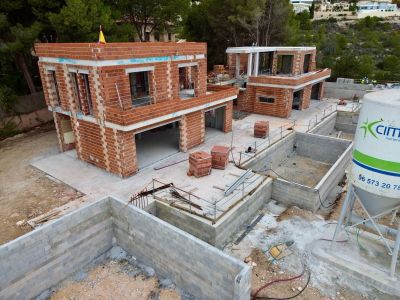 Aerial view of a villa project in its construction phase, featuring the brickwork of two main structures and concrete block foundations, set on a green hillside.