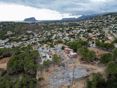 Aerial view of villas under construction within a residential area of Moraira, showing cranes and foundations, framed by the sea and Peñón de Ifach in the distance.