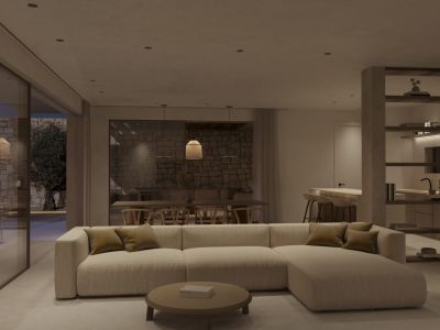 Open-plan living and dining area featuring a modular sofa, round coffee table, and sliding doors to a stone-walled terrace with an olive tree. Integrated kitchen visible.