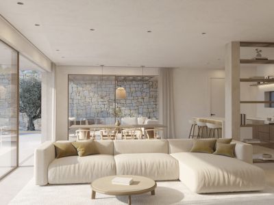 Spacious living area featuring a light sectional sofa and round coffee table. Large glass doors reveal an exterior patio with a natural stone wall and an ancient olive tree.