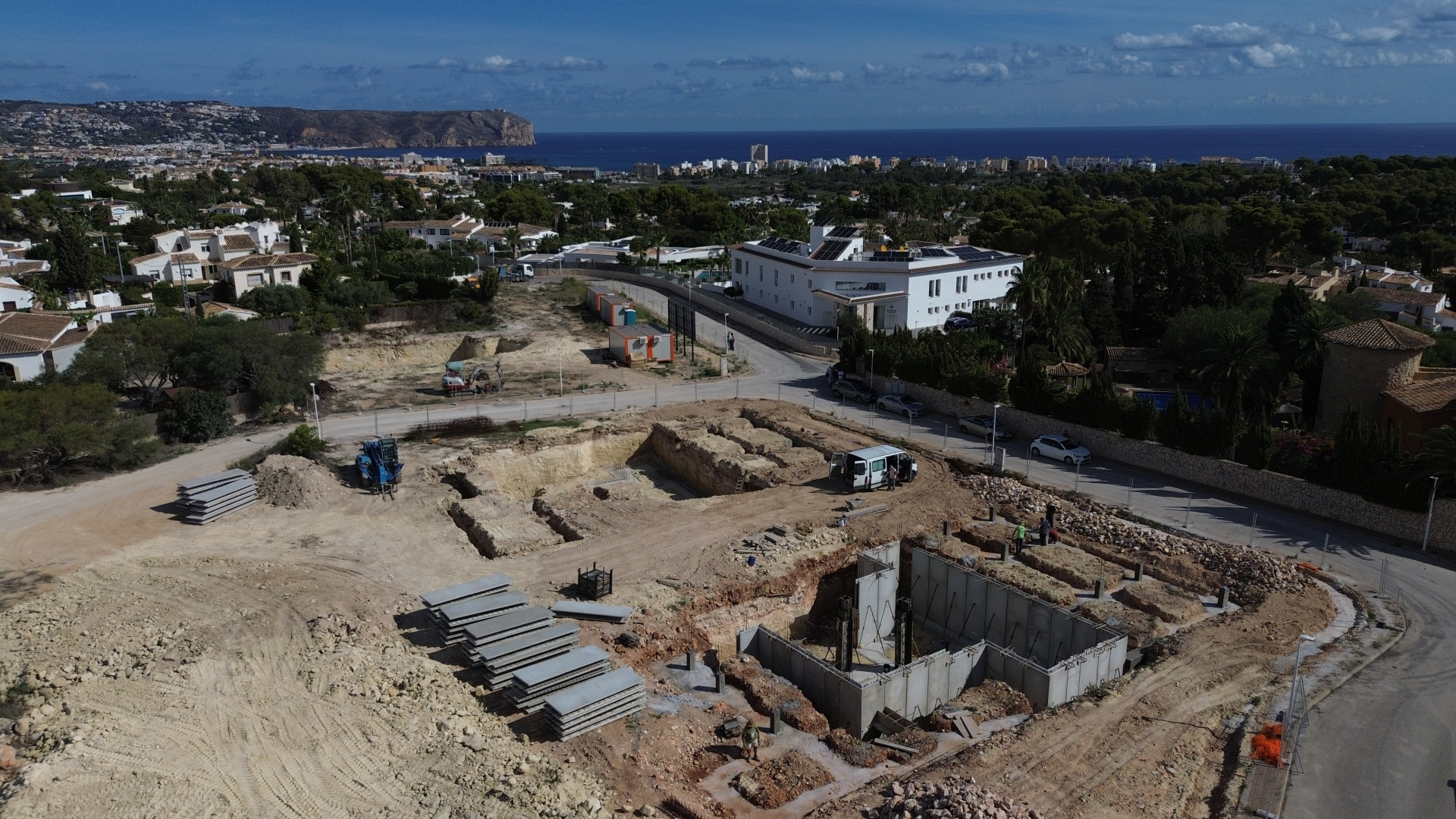 Aerial view of the construction site showing concrete foundations and ground excavations. Jávea town, the Mediterranean Sea, and the San Antonio Cape are visible in the background.