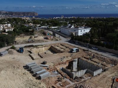 Aerial view of the construction site showing concrete foundations and ground excavations. Jávea town, the Mediterranean Sea, and the San Antonio Cape are visible in the background.