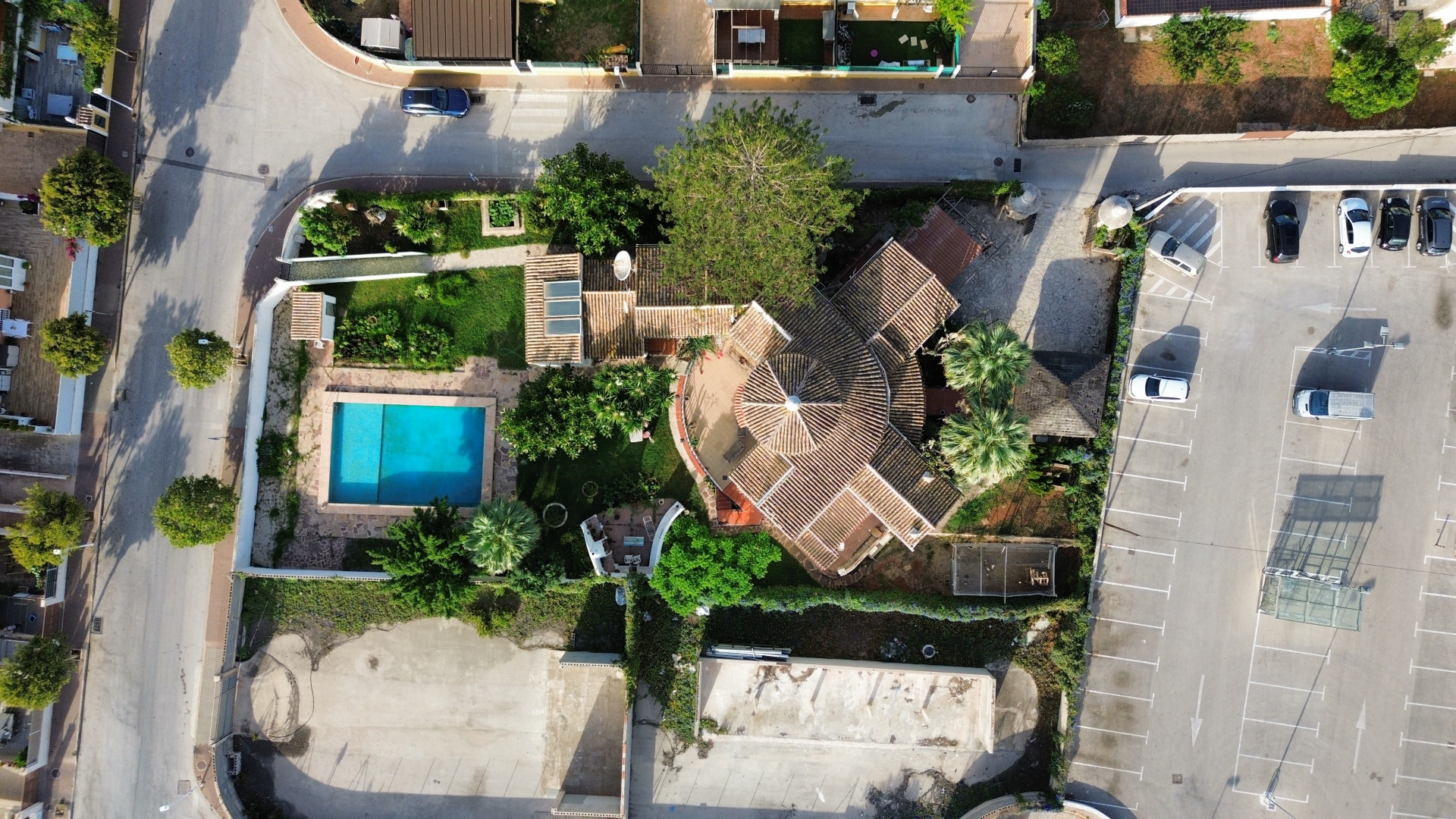 Aerial view of villa with pool and tiled roof, surrounded by greenery and adjacent parking lot.