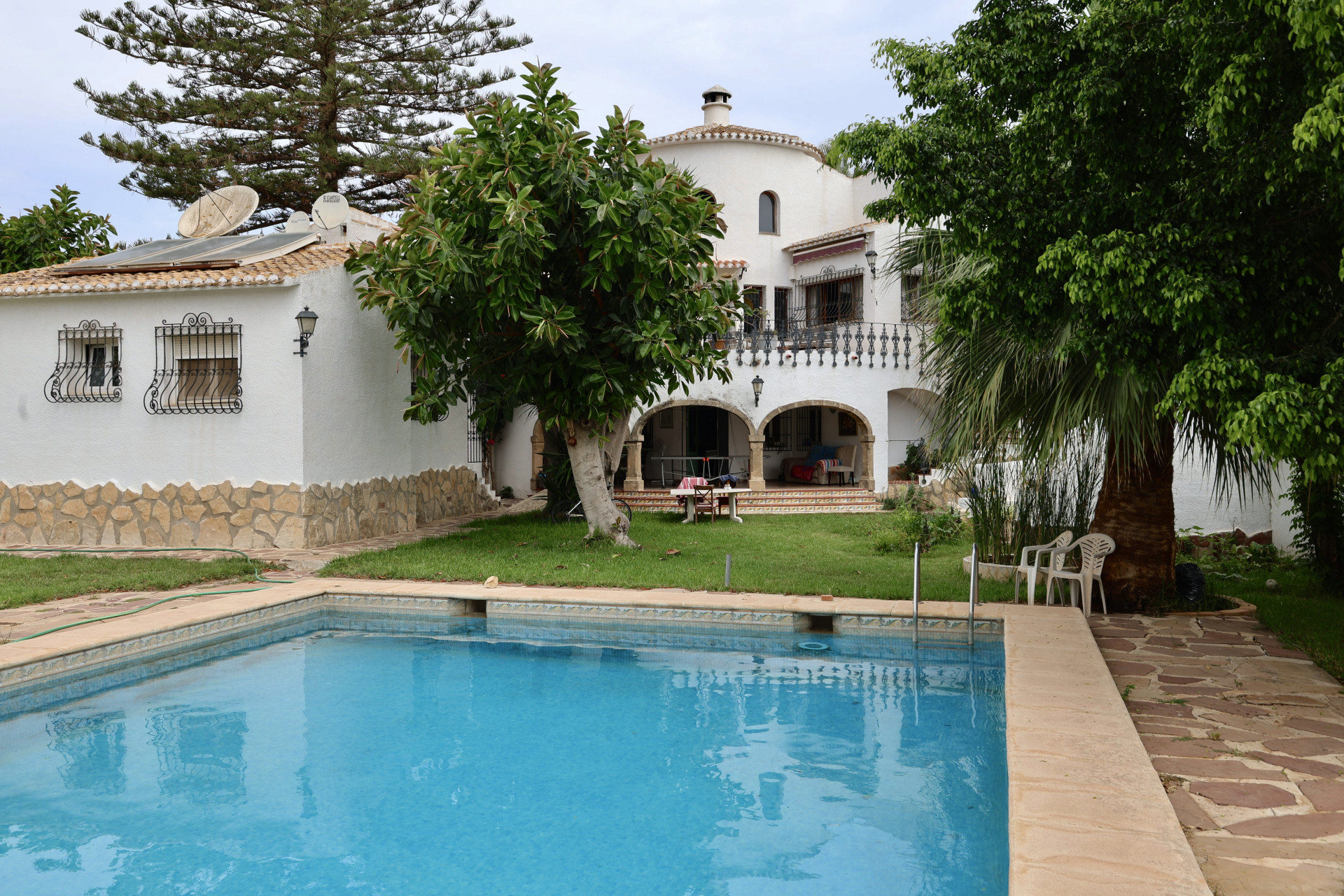 View of the turquoise blue pool and the villa's white facade with stone details and garden.