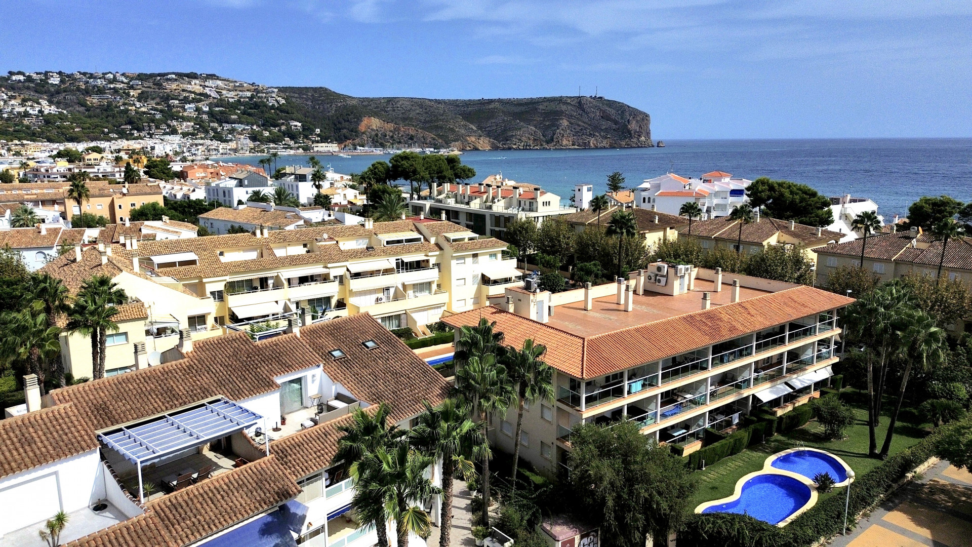 Aerial view of the urbanization with pool and red tile roofs, with the sea and coastline in the background.