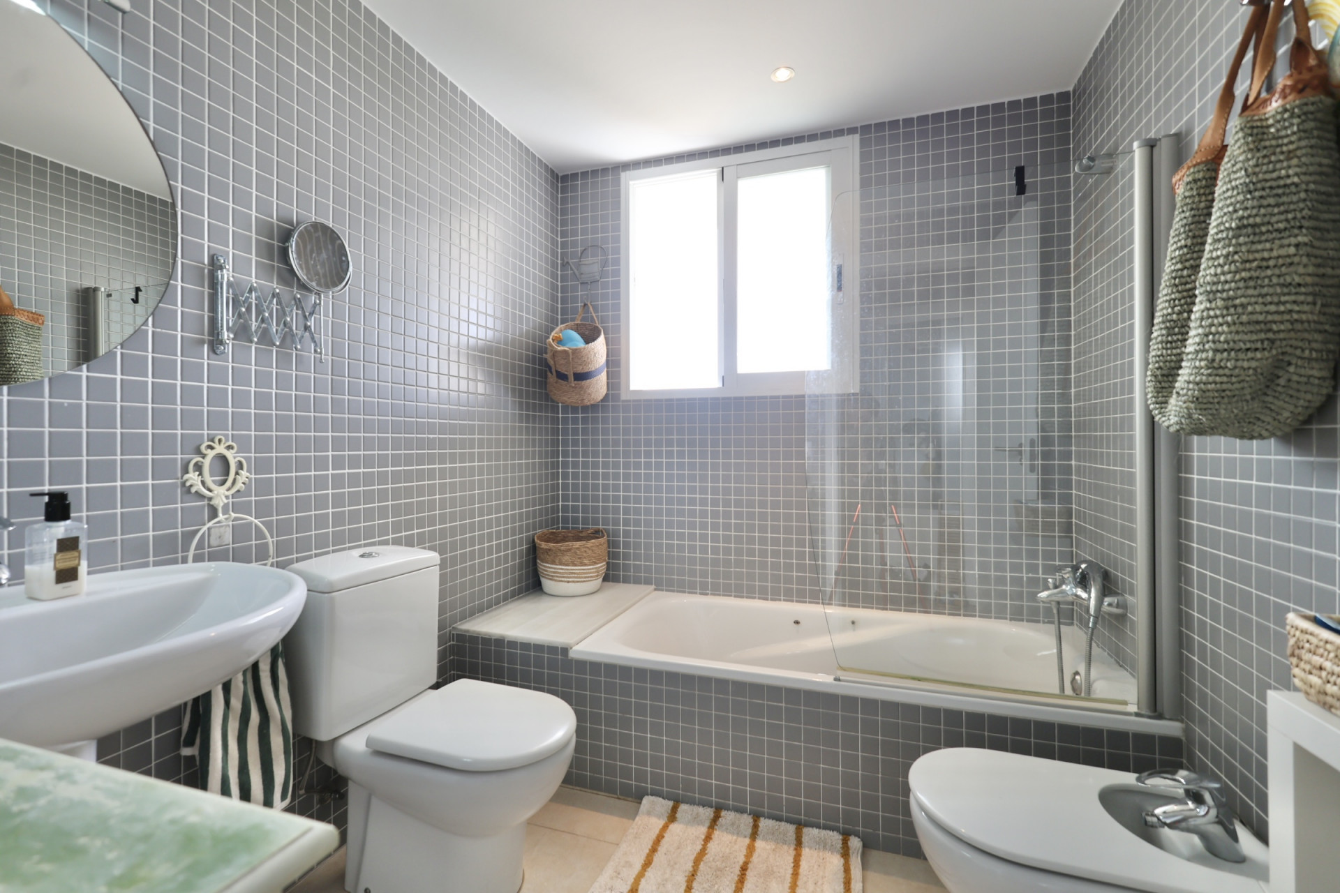 Bathroom featuring gray square tiling, pedestal sink, toilet, and bidet. A white bathtub sits beneath a window.