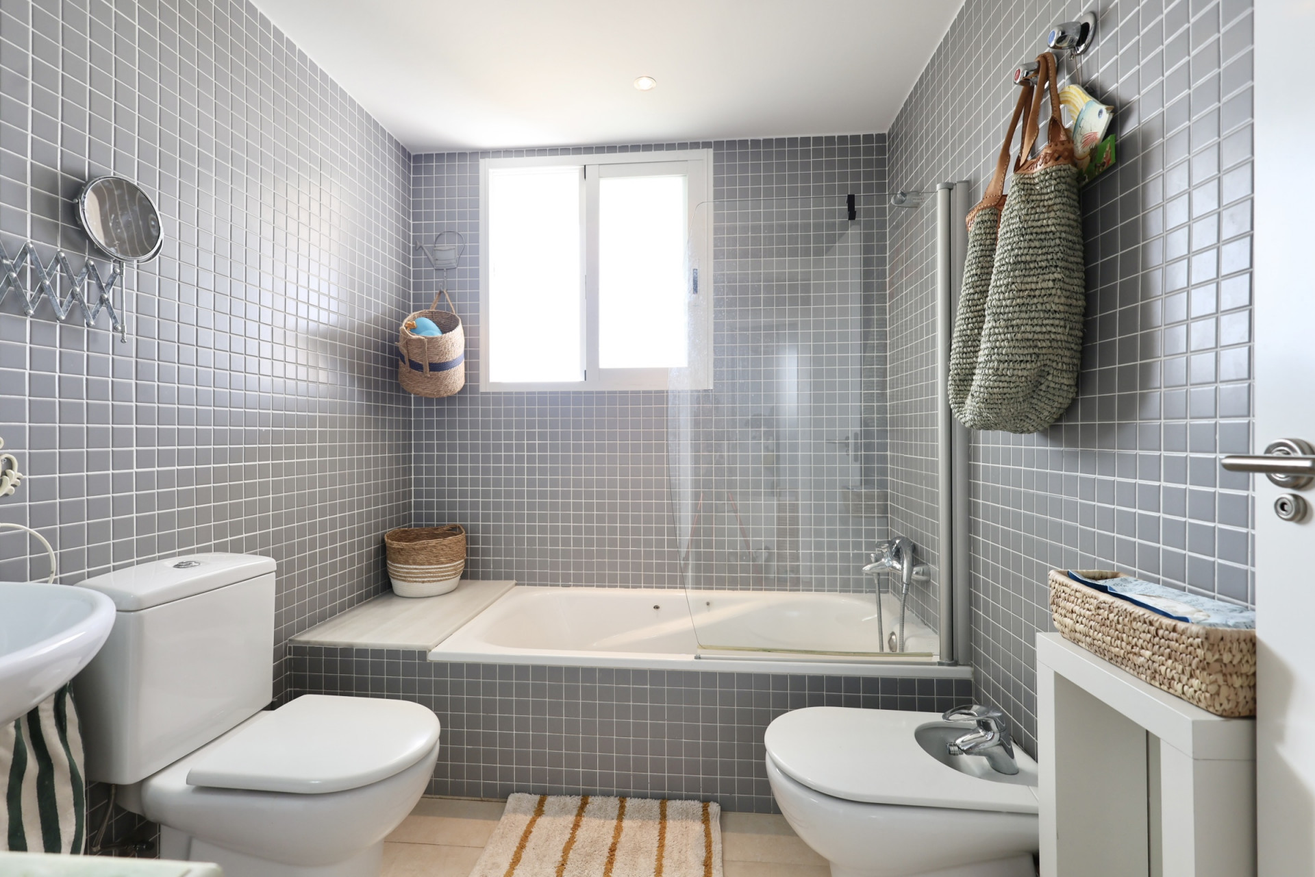 Bathroom featuring gray tiled walls, a bathtub with a glass screen, and wicker decorative elements.