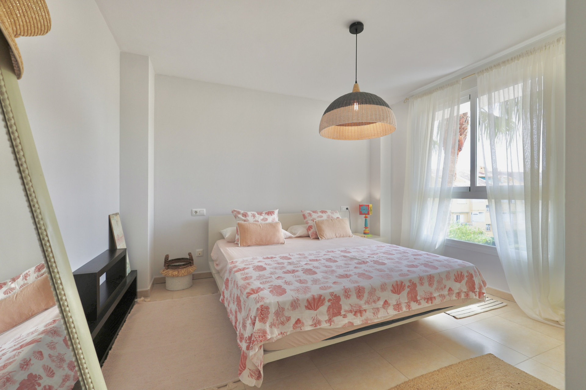 Bright bedroom featuring a double bed, patterned bedding, and a wicker pendant lamp.