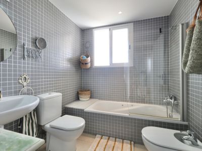 Bathroom featuring gray square tiling, pedestal sink, toilet, and bidet. A white bathtub sits beneath a window.