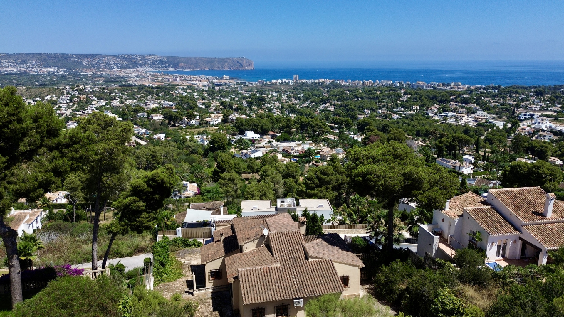 Aerial view of Jávea, featuring traditional villas with terracotta roofs nestled among pine trees, with the Mediterranean Sea and Cabo de San Antonio in the distance.