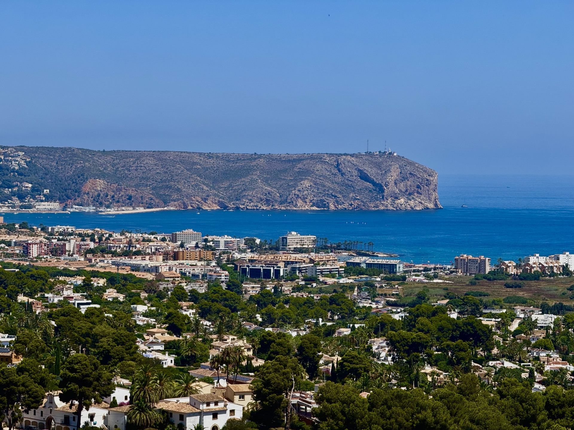 Expansive panoramic view showcasing Jávea's residential landscape, the deep blue Mediterranean Sea with boats, and the prominent rocky cape with its lighthouse.