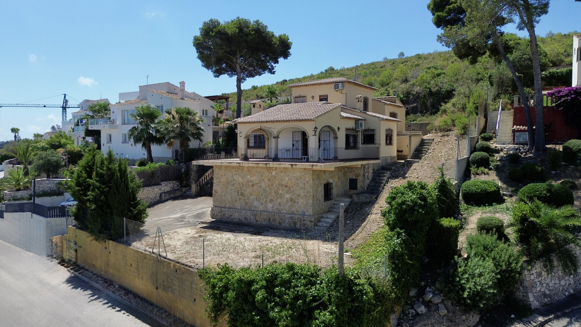 Exterior view of a two-story Mediterranean villa with a stone base and arched balcony. Situated on sloping terrain with sparse vegetation under a clear blue sky.