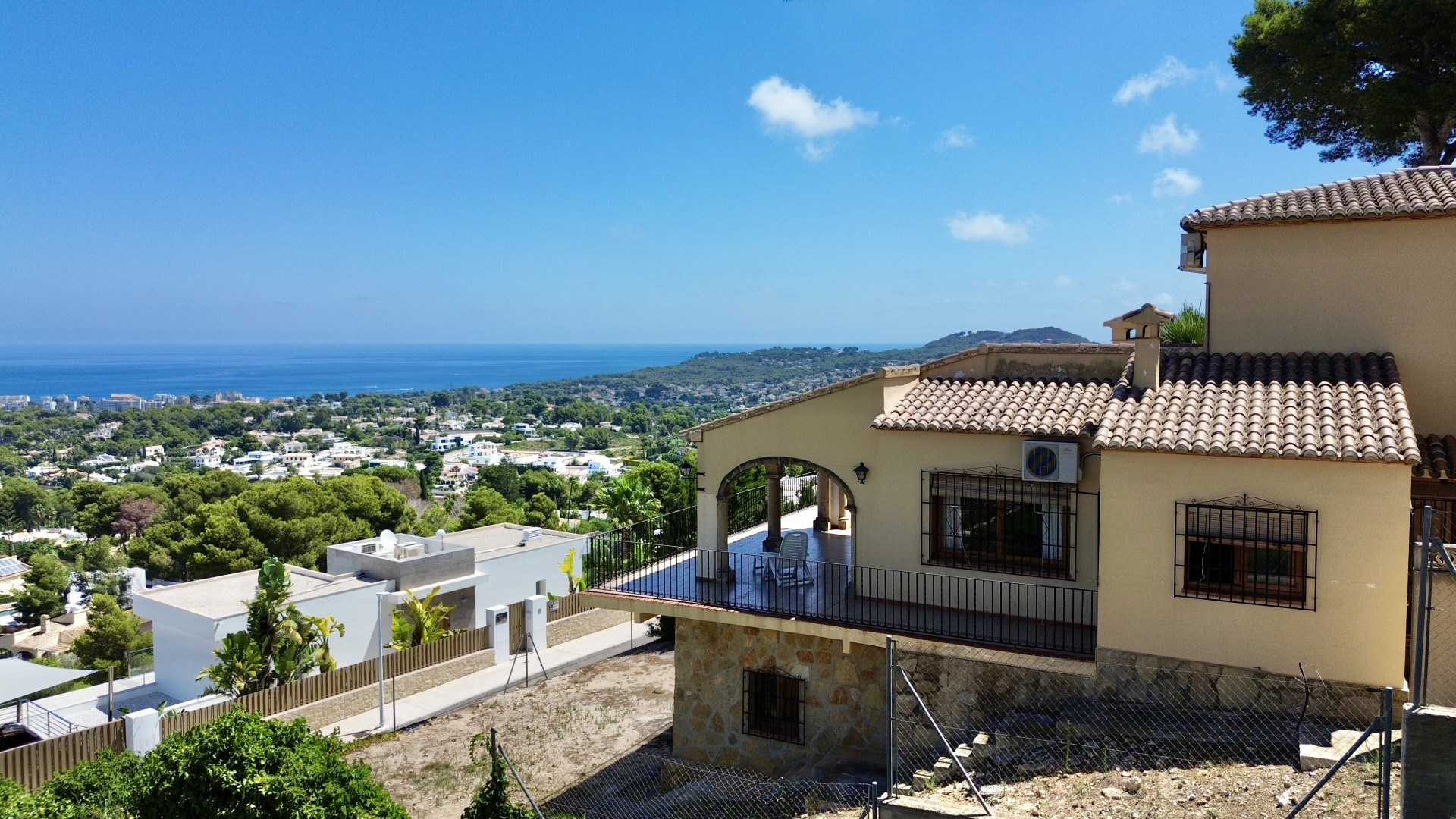 Exterior view of the villa featuring a covered terrace with arched design and black railings. Panoramic sea, town, and mountain views are visible from the sloping plot.