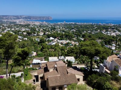 Aerial view of Jávea, featuring traditional villas with terracotta roofs nestled among pine trees, with the Mediterranean Sea and Cabo de San Antonio in the distance.