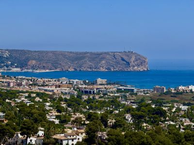 Expansive panoramic view showcasing Jávea's residential landscape, the deep blue Mediterranean Sea with boats, and the prominent rocky cape with its lighthouse.