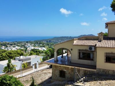 Exterior view of the villa featuring a covered terrace with arched design and black railings. Panoramic sea, town, and mountain views are visible from the sloping plot.