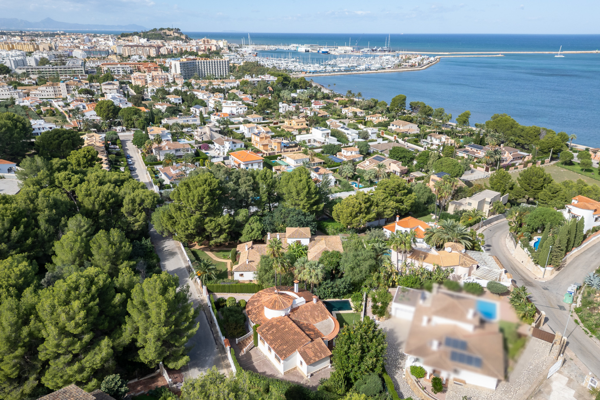 Aerial view showcasing the villa with its distinct red-tiled roof and private pool, nestled within a green residential area of Dénia, overlooking the marina and sea.