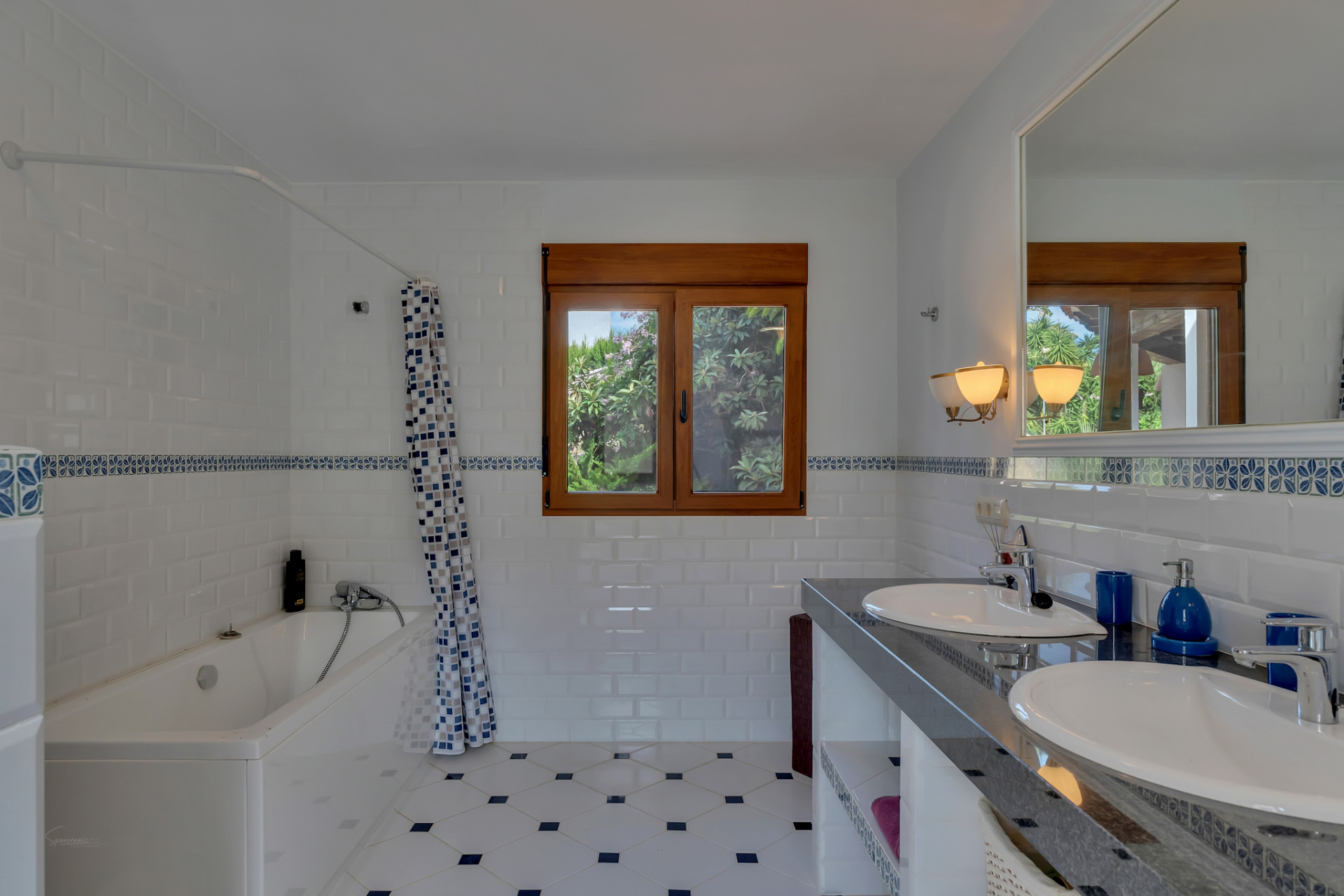 Bathroom featuring a double vanity with dark countertop, two sinks, and a large mirror. White subway tiles with a blue decorative border complement the patterned floor and wooden window frame overlooking greenery.