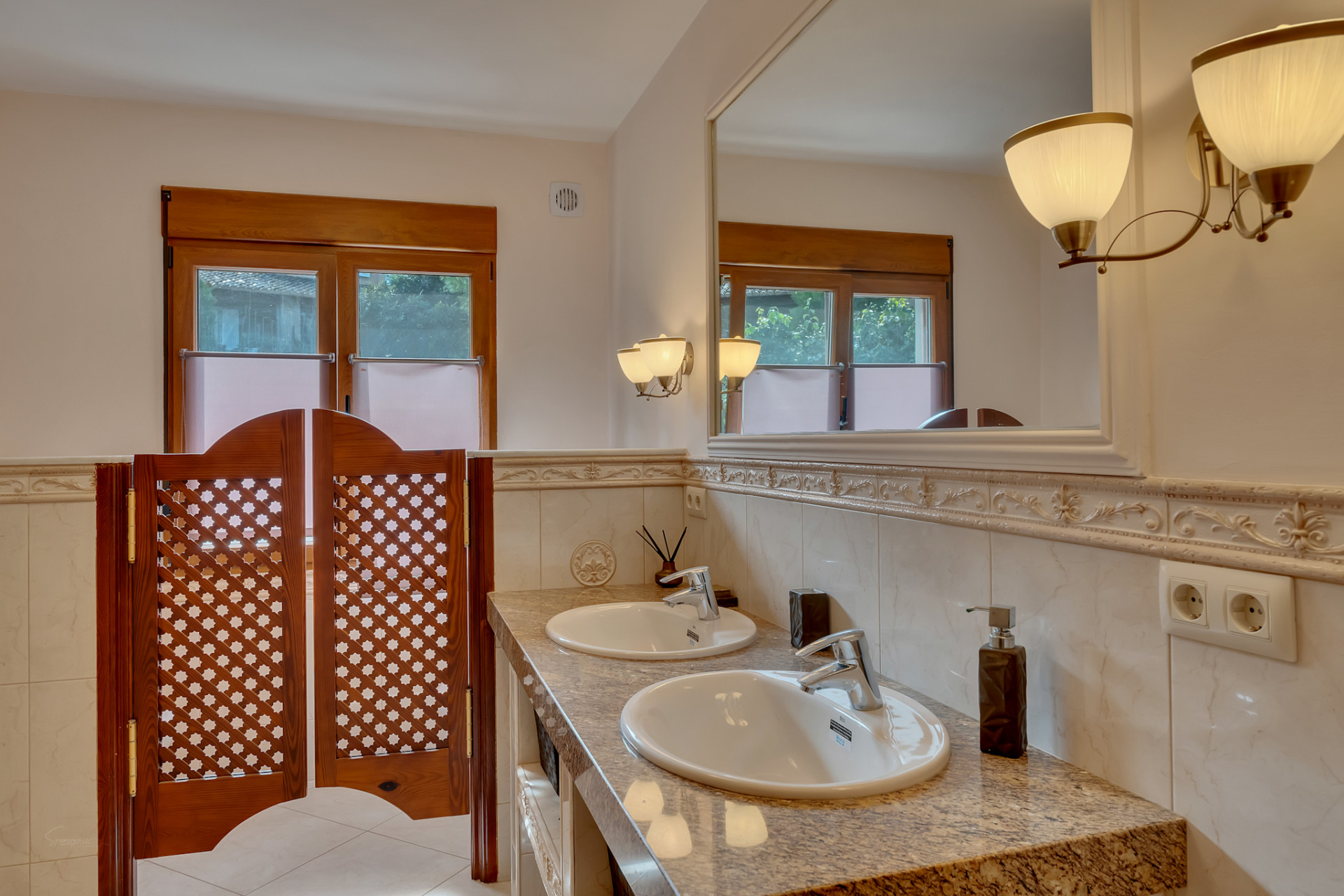 Bathroom with double granite vanity, two sinks, and a large framed mirror with elegant wall sconces. Features wooden windows and lattice saloon doors.