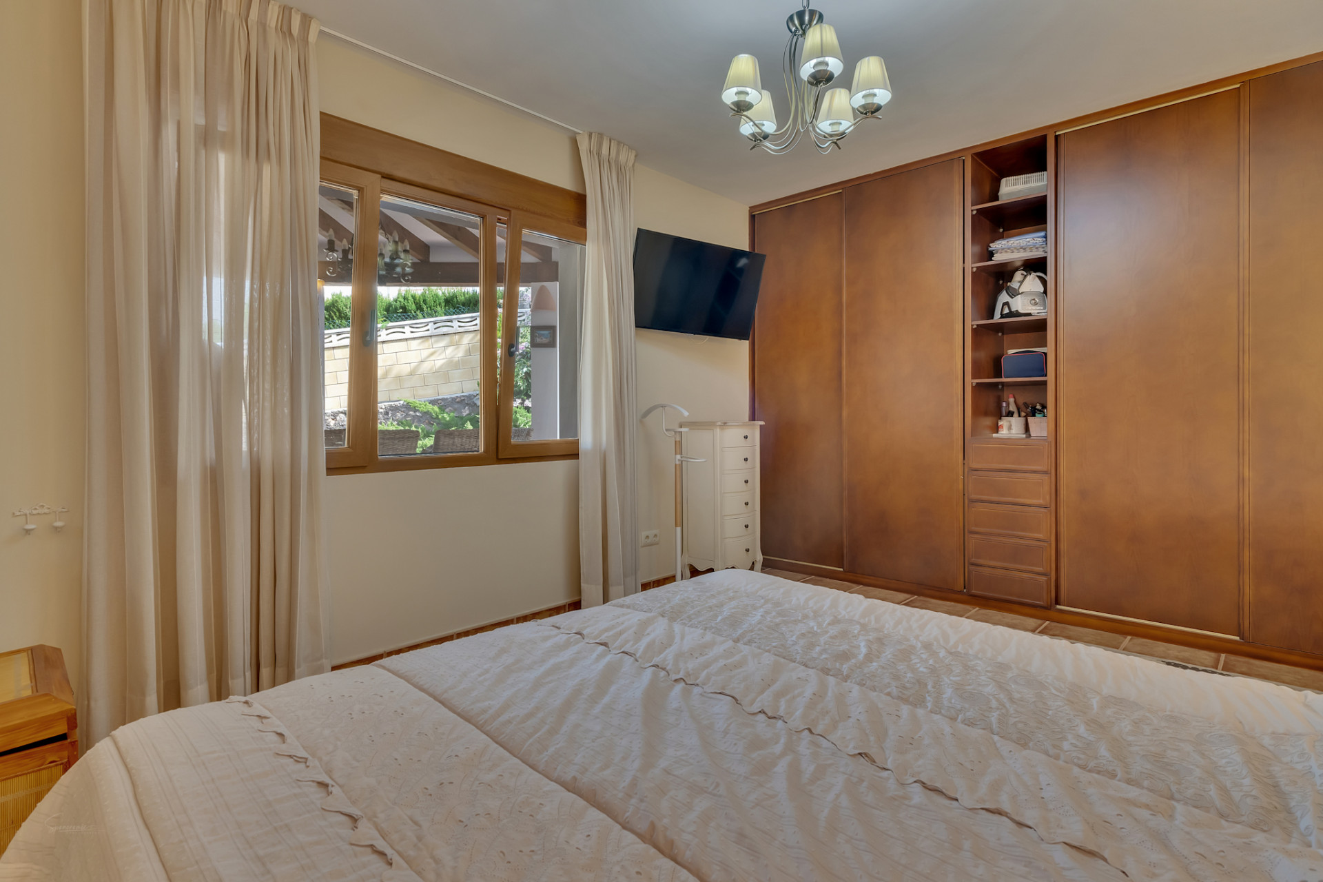 Bedroom featuring a double bed, integrated wooden wardrobes, and a window overlooking a landscaped wall. Includes a wall-mounted TV and white dresser.