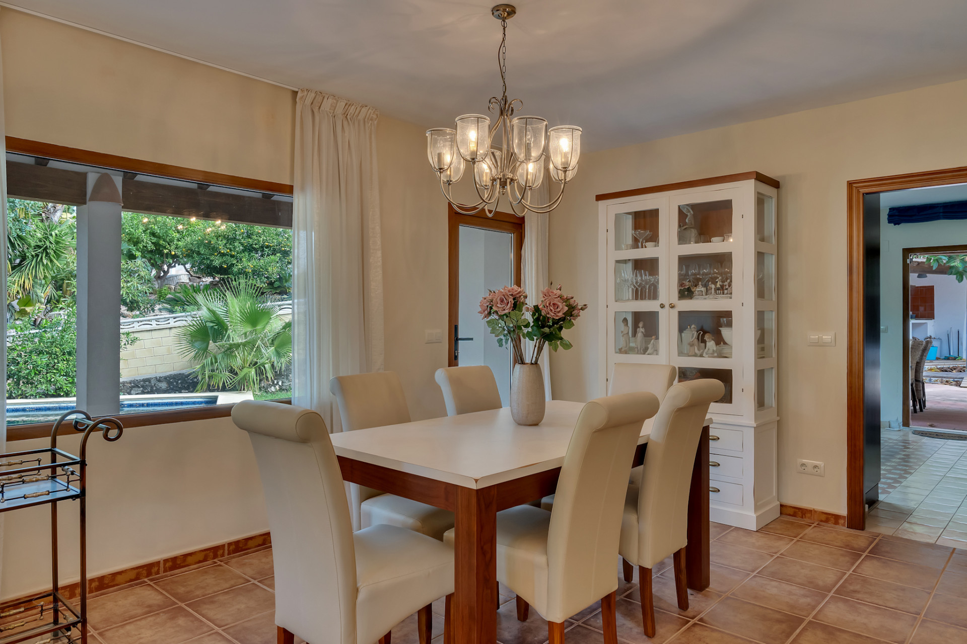 Bright dining room with a wooden table, cream upholstered chairs, and a chandelier. A large window offers views of the lush garden and private pool.