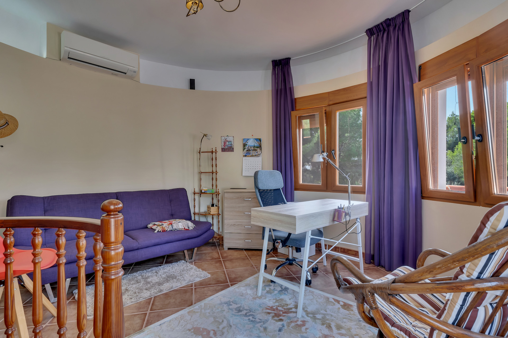 Bright office space featuring a purple sofa, light wood desk with an ergonomic chair, and large wooden windows offering views of lush greenery. Includes an air conditioning unit and terracotta floor tiles.