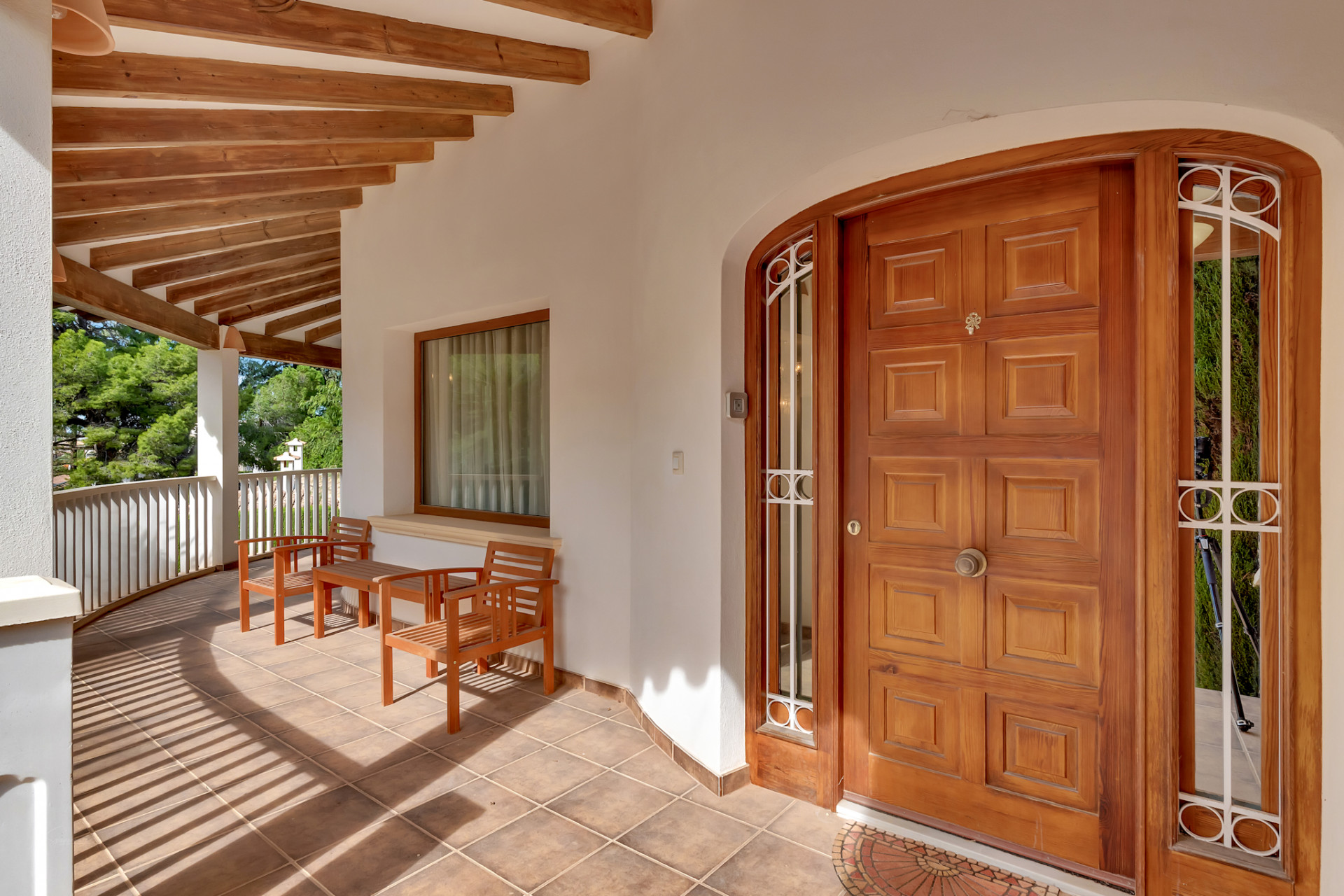 Covered entrance terrace with a paneled wooden door, tiled floor, and exposed wooden beams. Features a seating area and a curved railing overlooking lush greenery.