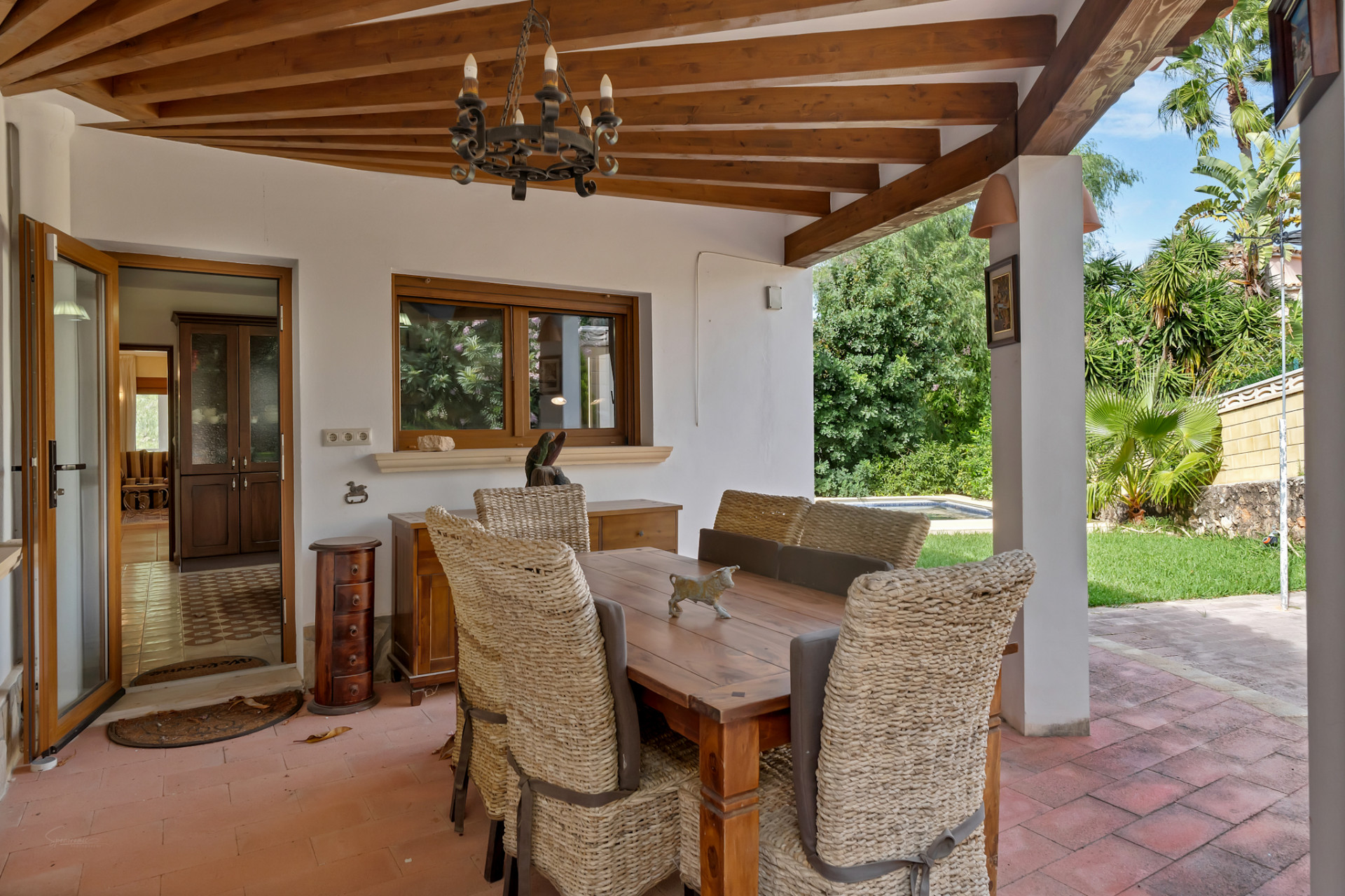 Covered outdoor dining area with a wooden beamed ceiling, terracotta floor, and a rustic chandelier. Wicker chairs surround a robust wooden table, opening to a lush garden and interior.