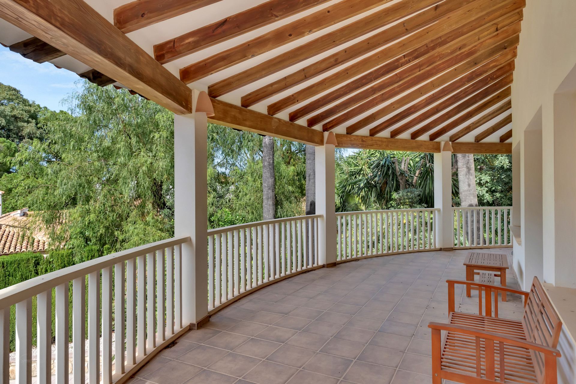 Curved covered terrace featuring a tiled floor and an exposed wooden beam ceiling. A white balustrade frames views of lush greenery, complemented by simple wooden seating.