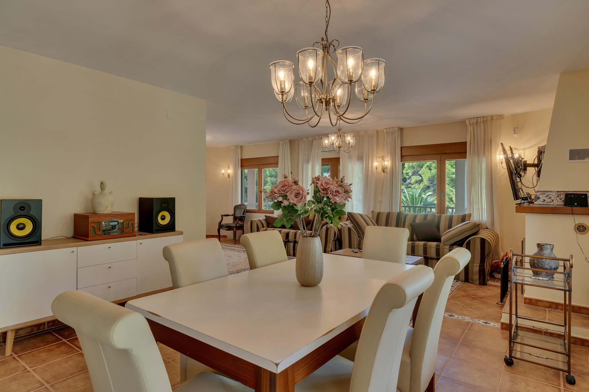 Elegant dining area featuring a classic chandelier and cream-colored chairs, seamlessly connecting to a bright living space with striped sofas and large windows overlooking lush greenery.